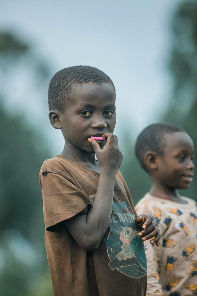 Two African boys standing outside, one looking thoughtfully with a pencil in hand.